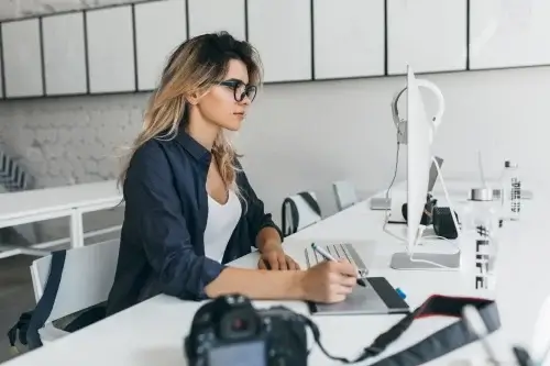 stylish-fair-haired-girl-using-tablet-after-photoshoot-sitting-table-with-computer-camera-charming-female-student-glasses-black-shirt-working-project500